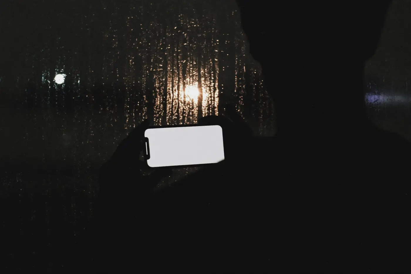 Sylwetka osoby z telefonem przy oknie zalanym deszczem, nocą, z rozmytymi światłami miasta